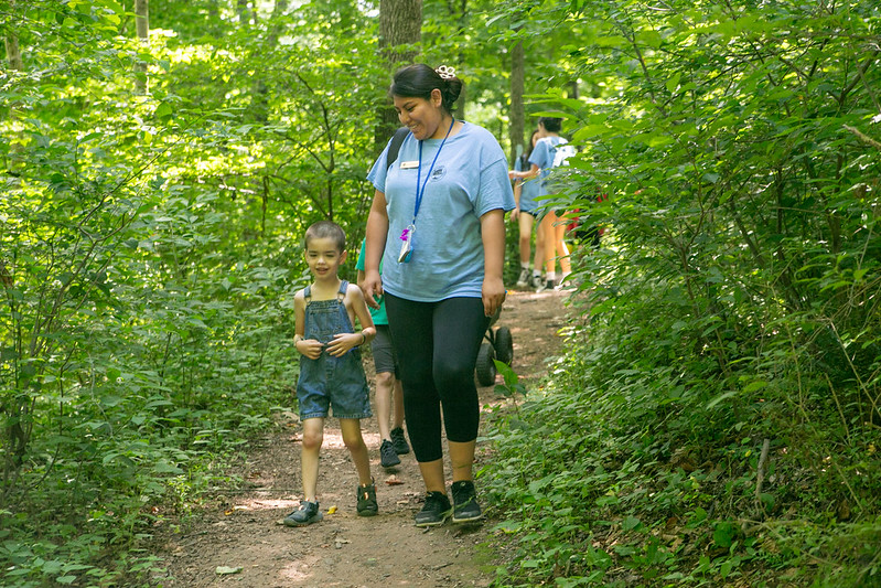 Program Access inclusion support staff walking next to camper down a natural surface trail.