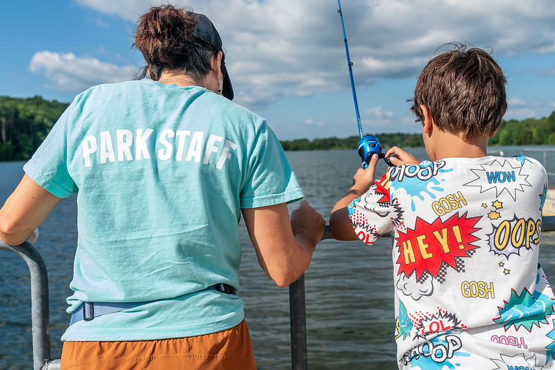 Park staff standing next to program participant with fishing rod in the water waiting to catch a fish during inclusive fishing program.