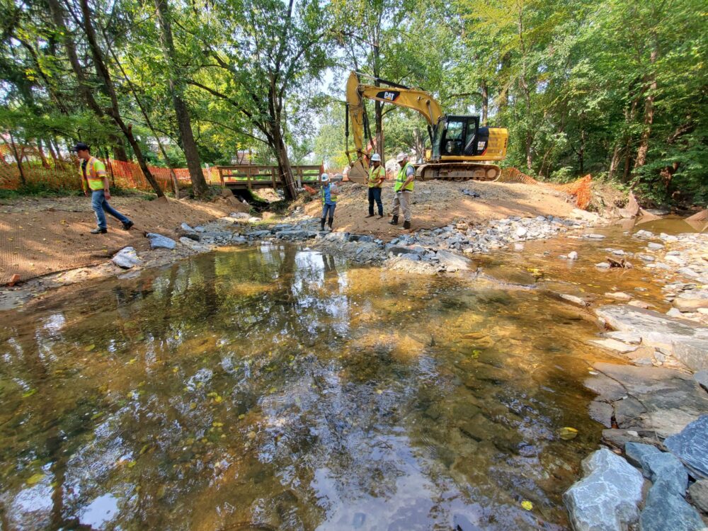 A stream with construction equipment, orange construction fencing, and staff in yellow vests in the background.