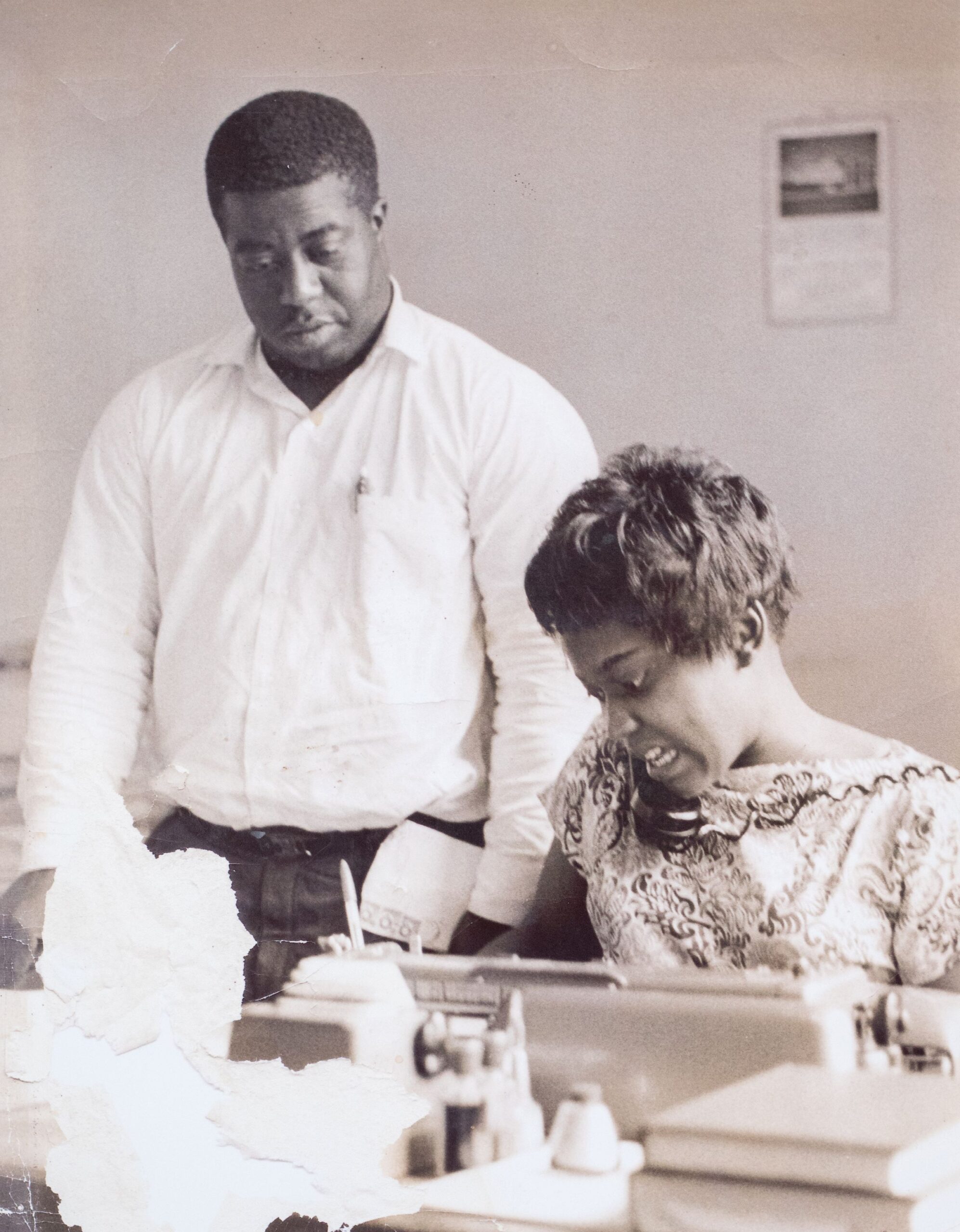 A black and white photo shows an African American man standing next to a seated African American woman at a typewriter.