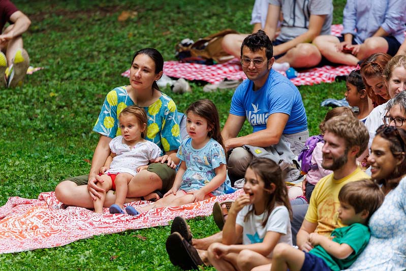 family sitting in the grass watching Happenstance Theater production in summer of 2023